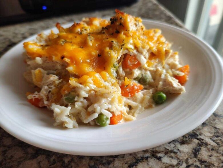 A close-up serving of creamy rice casserole topped with melted cheddar cheese, peas, and carrots on a white plate.