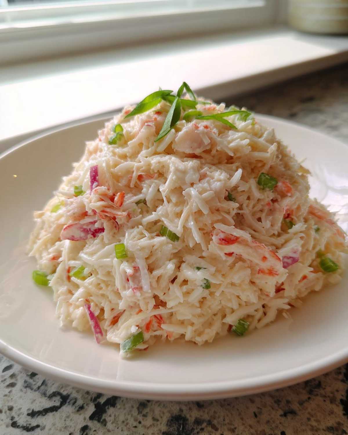 A mound of creamy seafood salad imitation crab mixed with red onion and green onion, garnished on a white plate.