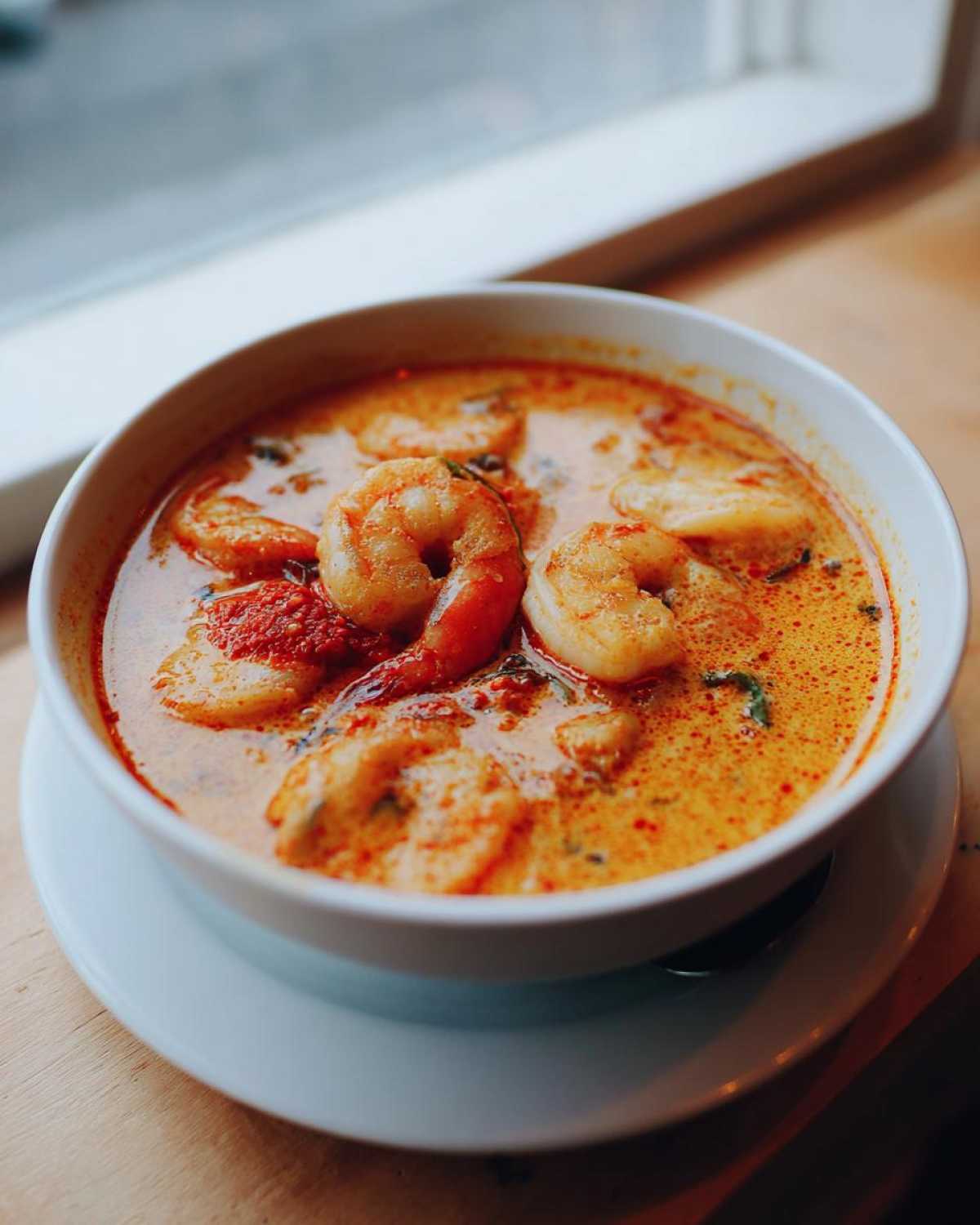 Close-up of a white bowl filled with creamy, orange-hued tom yum soup featuring large shrimp.