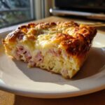 A golden-brown slice of Croissant Breakfast Casserole featuring visible chunks of ham and melted cheese on top.