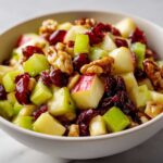 A close-up view of a vibrant apple salad featuring diced apples, celery, walnuts, and dried cranberries in a white bowl.