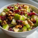 Close-up of a white bowl filled with crunchy apple salad featuring diced green apples, celery, walnuts, and dried cranberries.