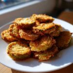 A pile of crispy, golden Delicious Air Fryer Fried Pickles stacked high on a white plate, illuminated by sunlight.