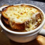 Close-up of a bowl of french onion soup topped with a thick slice of toasted bread and melted, bubbly Gruyère cheese.