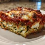 A close-up of a hearty slice of zucchini lasagna showing layers of ricotta, zucchini, and bubbling tomato sauce.