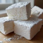 Close-up of four large, fluffy homemade marshmallows dusted heavily with powdered sugar on a wooden surface.