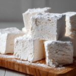 Close-up of several large, fluffy homemade marshmallows stacked on a wooden board, heavily dusted with powdered sugar.