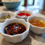 Four small white dishes holding different types of Dumpling Sauce, including chili oil, sweet chili, and green relish.