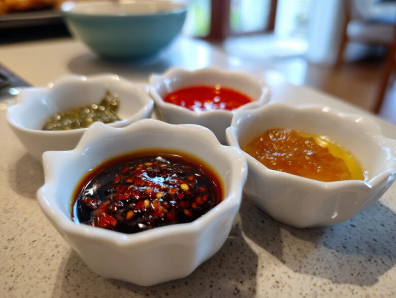 Four small white dishes holding different types of Dumpling Sauce, including chili oil, sweet chili, and green relish.