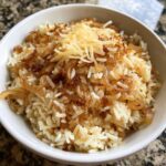 Close-up of a white bowl filled with french onion butter rice topped with caramelized onions and shredded cheese.