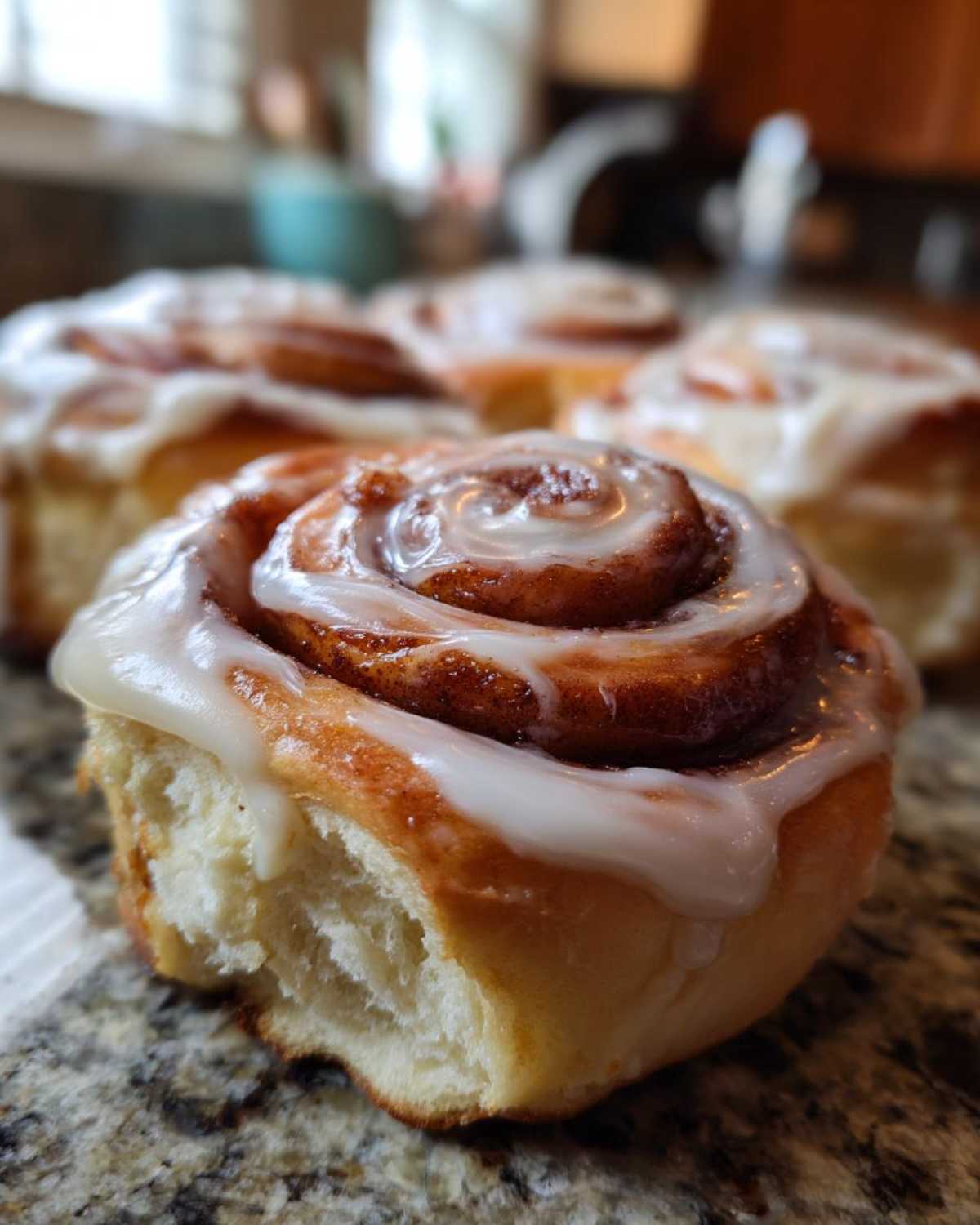 A close-up, low-angle shot of a soft, freshly baked cinnamon bun topped with white glaze, with more cinnamon buns blurred in the background.