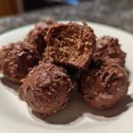 Close-up of several chocolate-coated Frozen Peanut Butter Greek Yogurt Bites on a white plate, one is cut open.