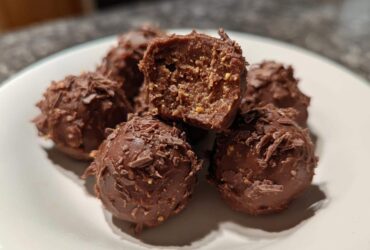 Close-up of several chocolate-coated Frozen Peanut Butter Greek Yogurt Bites on a white plate, one is cut open.