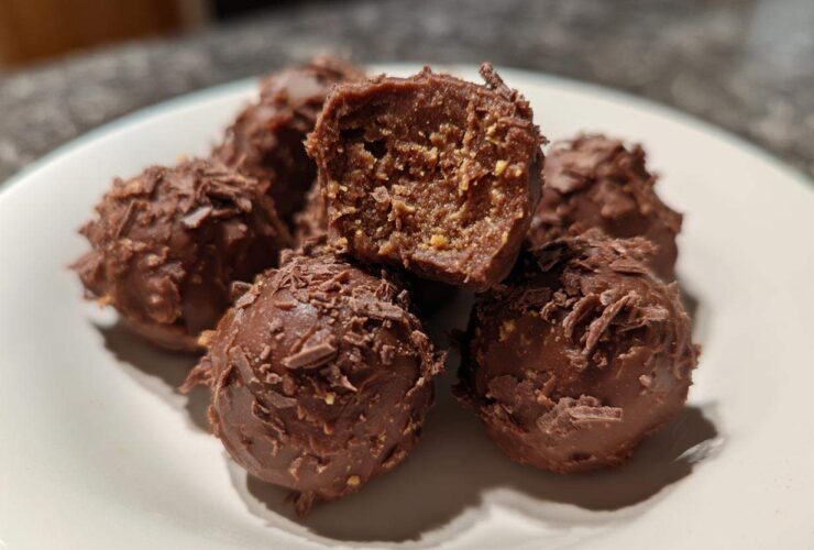 Close-up of several chocolate-coated Frozen Peanut Butter Greek Yogurt Bites on a white plate, one is cut open.