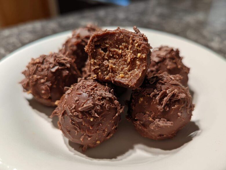 Close-up of several chocolate-coated Frozen Peanut Butter Greek Yogurt Bites on a white plate, one is cut open.