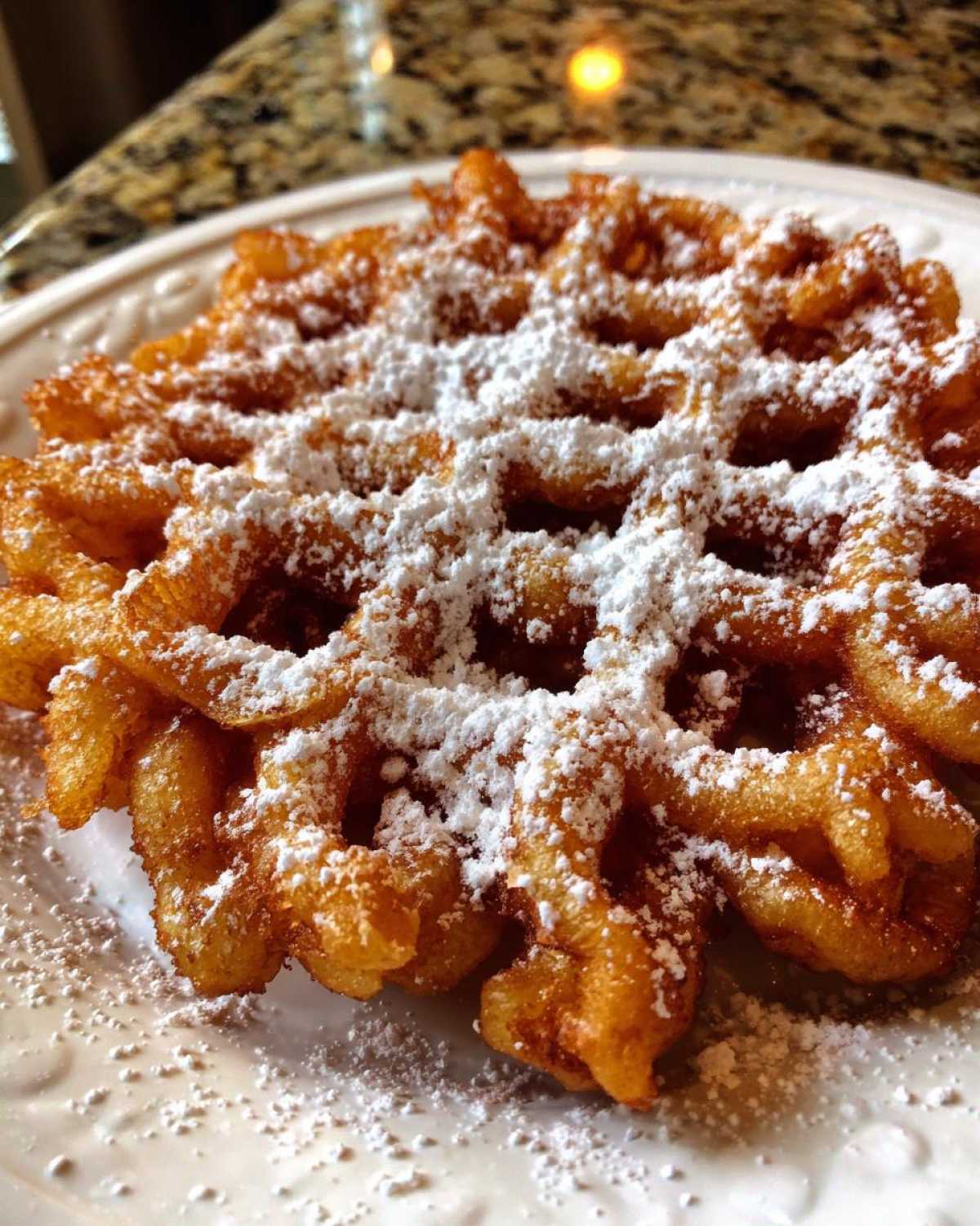 A golden brown, freshly fried funnel cake recipe dessert generously dusted with powdered sugar on a white plate.
