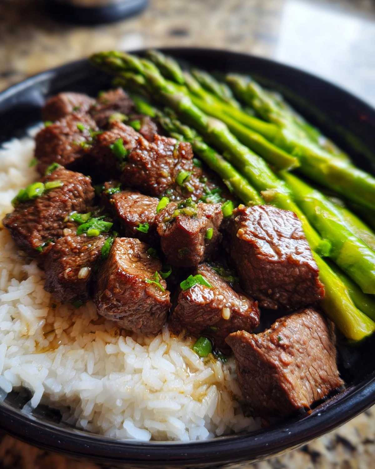 Close-up of a Garlic Butter Steak Bowl with rice, cubed steak coated in sauce, and bright green asparagus spears.
