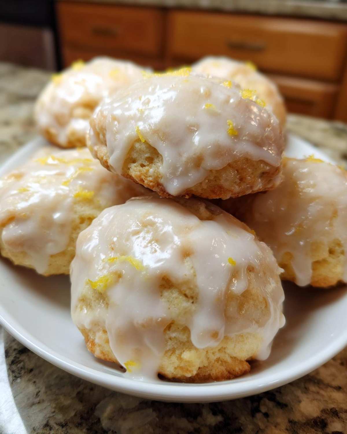 A close-up of soft Italian ricotta cookies piled on a white plate, topped with thick white glaze and bright yellow lemon zest.