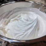 Close-up of thick, glossy white royal icing recipe mixture whipped to stiff peaks in a stainless steel bowl.