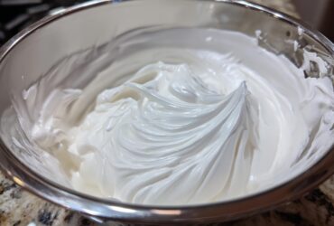 Close-up of thick, glossy white royal icing recipe mixture whipped to stiff peaks in a stainless steel bowl.