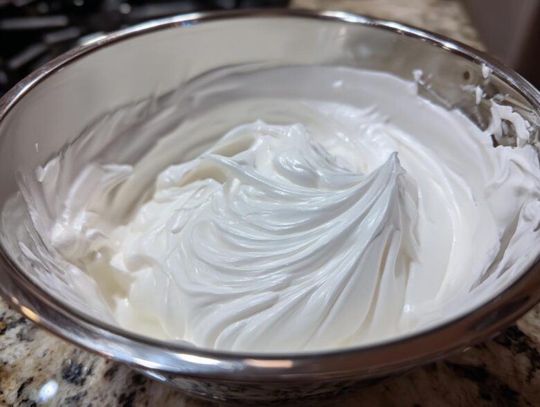 Close-up of thick, glossy white royal icing recipe mixture whipped to stiff peaks in a stainless steel bowl.