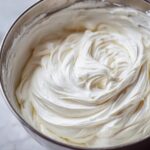 Close-up of thick, glossy white royal icing recipe mixture swirled inside a stainless steel mixing bowl.