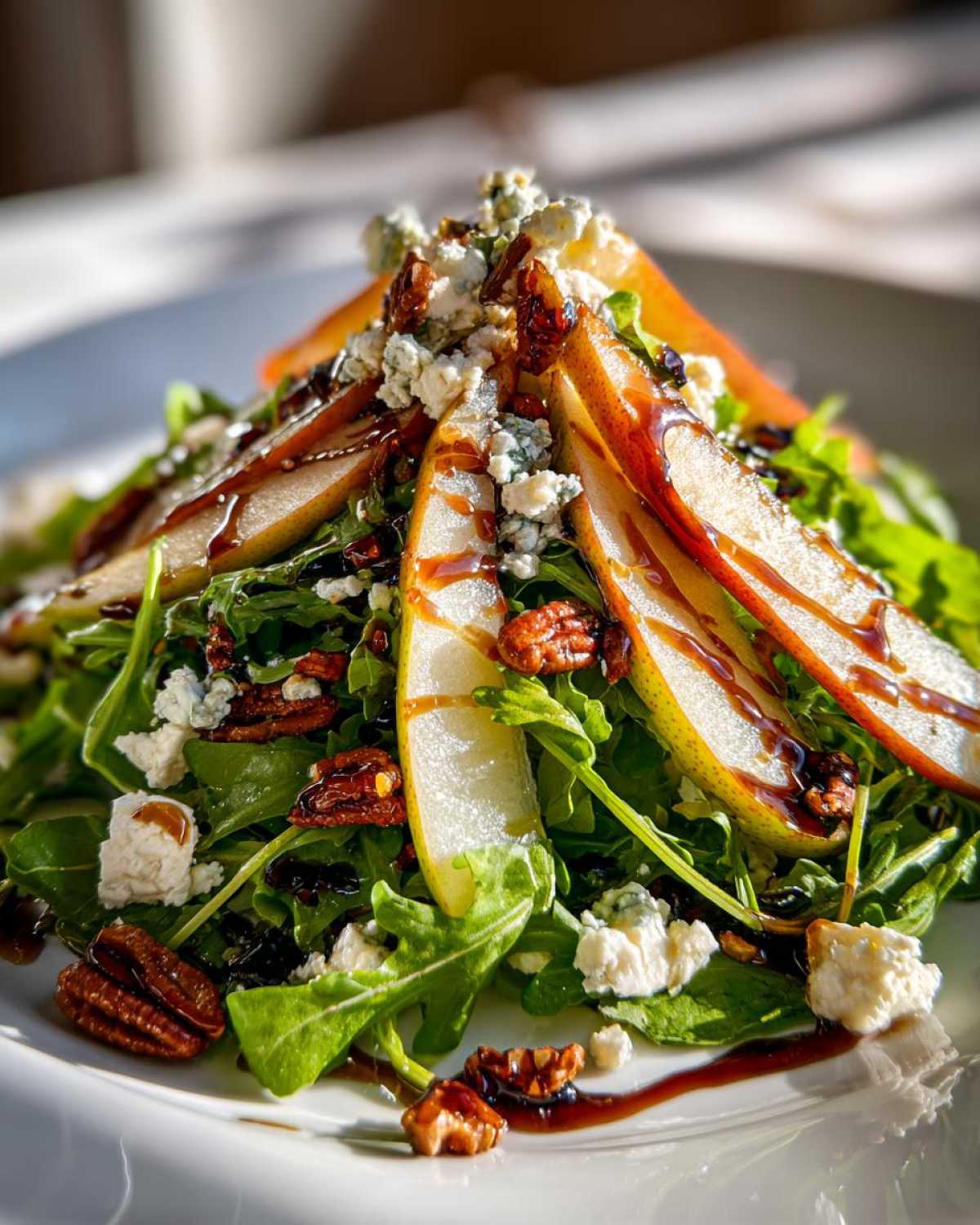 A vibrant close-up of a gourmet pear salad featuring arugula, sliced pears, blue cheese crumbles, candied pecans, and a balsamic glaze.