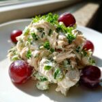 A mound of creamy greek yogurt chicken salad garnished with fresh parsley and surrounded by red grapes on a white plate.