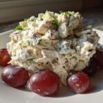 A mound of creamy greek yogurt chicken salad garnished with herbs and served with red grapes on a white plate.