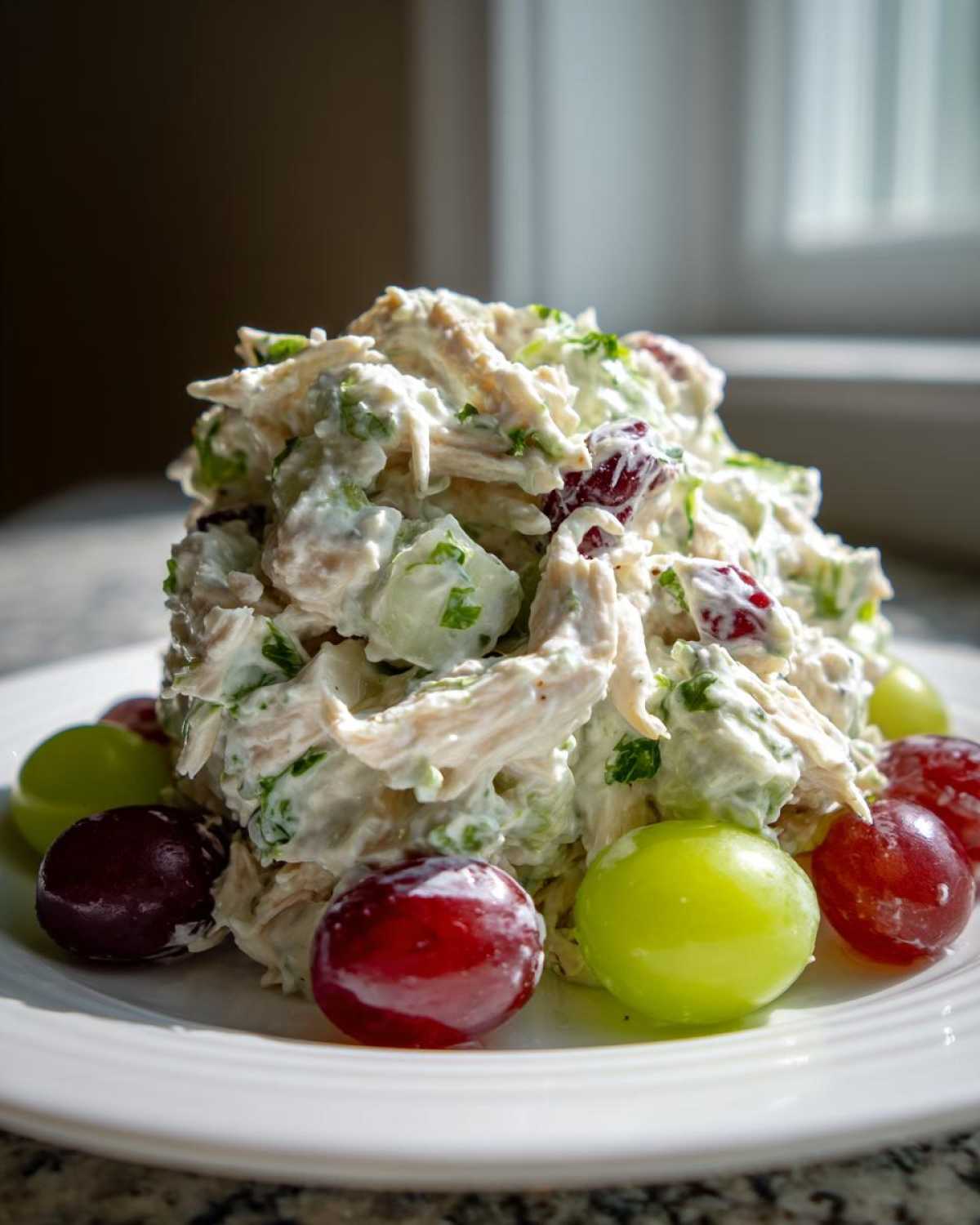 A mound of creamy greek yogurt chicken salad mixed with celery and cranberries, garnished with red and green grapes on a white plate.