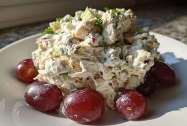 A mound of creamy greek yogurt chicken salad garnished with herbs and served with red grapes on a white plate.