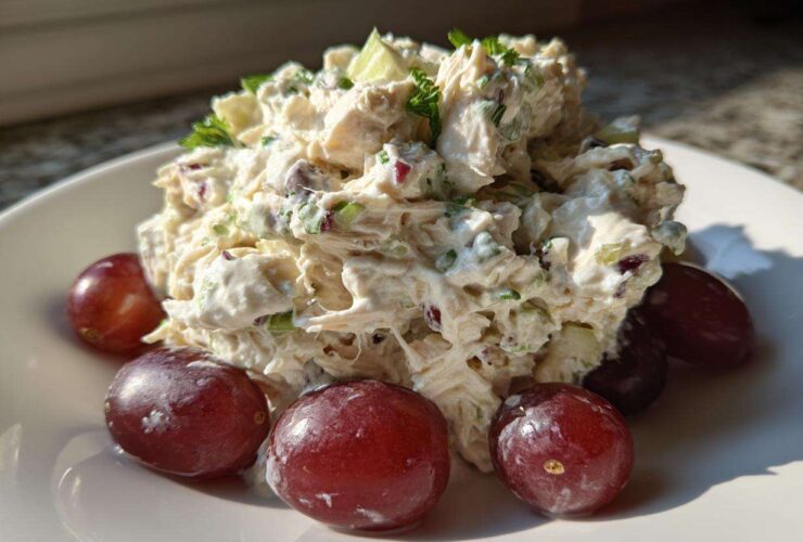 A mound of creamy greek yogurt chicken salad garnished with herbs and served with red grapes on a white plate.