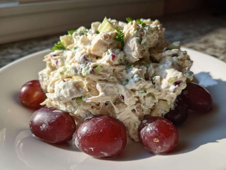 A mound of creamy greek yogurt chicken salad garnished with herbs and served with red grapes on a white plate.