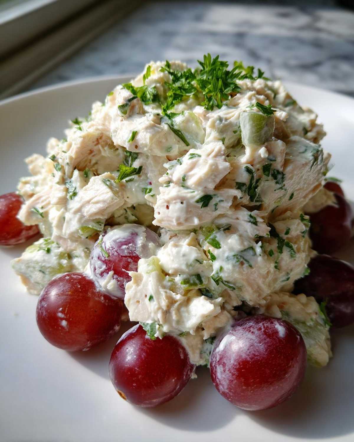 A mound of creamy greek yogurt chicken salad garnished with parsley, surrounded by fresh red grapes on a white plate.