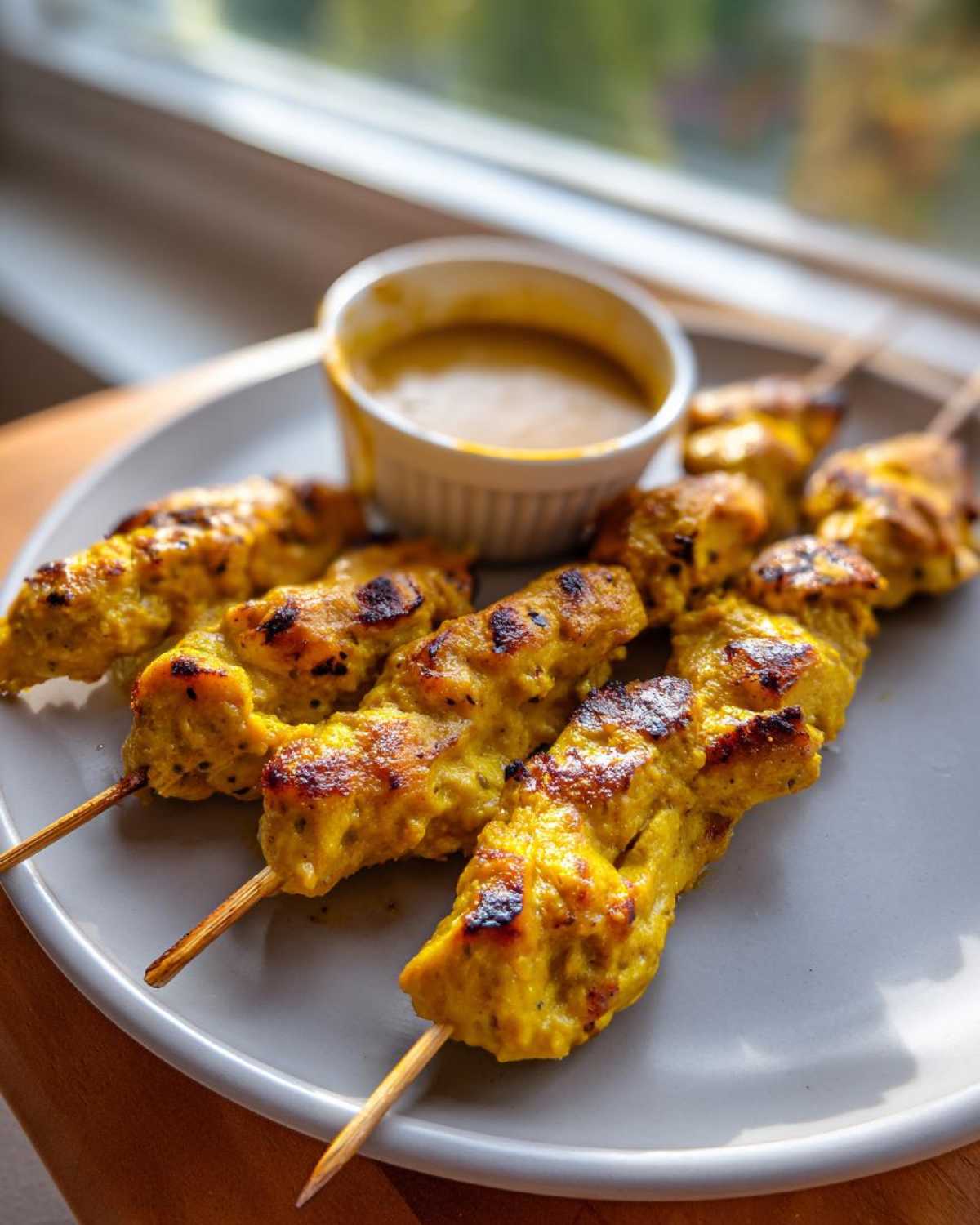 Close-up of grilled, yellow-marinated chicken satay skewers served on a plate next to a small bowl of peanut sauce.