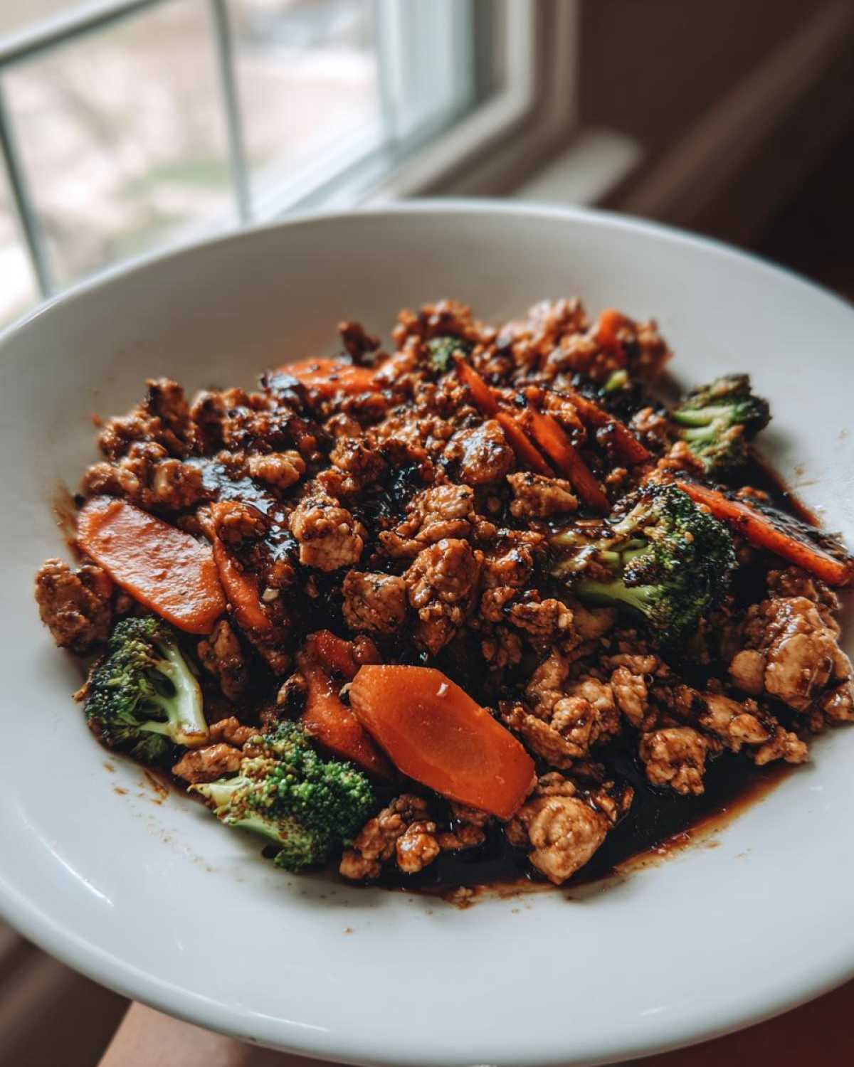 A close-up of a white bowl filled with Ground Chicken Teriyaki Stir Fry, featuring ground chicken, bright orange carrots, and green broccoli florets coated in dark sauce.