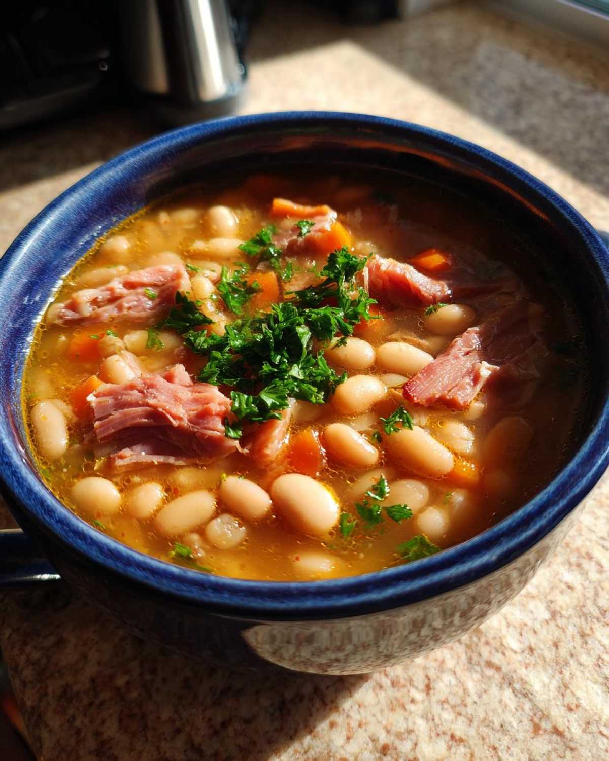 Close-up of a deep blue bowl filled with hearty navy bean soup, featuring white beans, chunks of ham, carrots, and fresh parsley garnish.