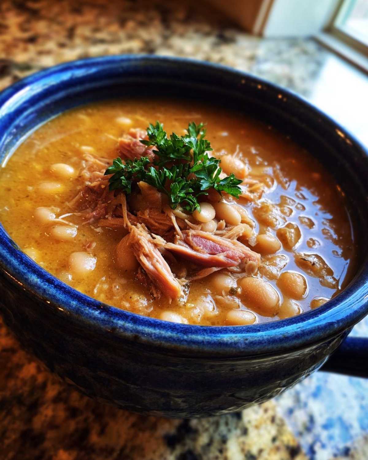 Close-up of a hearty navy bean soup topped with shredded pork and fresh parsley in a blue bowl.