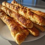 A pile of golden brown Homemade Breadsticks Inspired by Olive Garden stacked on a white plate.