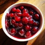 Close-up overhead shot of glistening, deep red cherry pie filling in a small white bowl, resting on a wooden surface.