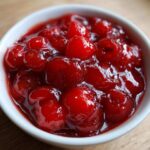A close-up view of glossy, bright red cherry pie filling packed with whole cherries in a small white bowl.