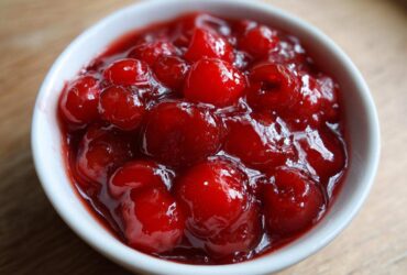 A close-up view of glossy, bright red cherry pie filling packed with whole cherries in a small white bowl.