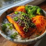 A close-up of a Honey Glazed Salmon Bowl featuring a glistening salmon fillet over rice, topped with sesame seeds and green onions.