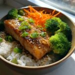 A close-up of a Honey Glazed Salmon Bowl featuring a glazed salmon fillet over white rice, served with bright green broccoli florets and shredded carrots.