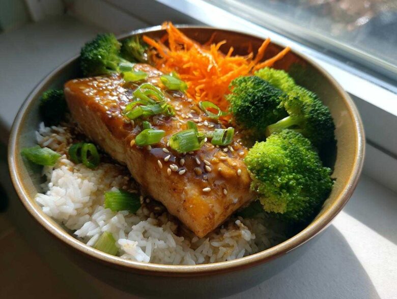 A close-up of a Honey Glazed Salmon Bowl featuring a glazed salmon fillet over white rice, served with bright green broccoli florets and shredded carrots.