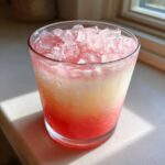 Close-up of a glass filled with Irresistible Strawberry Piña Colada Punch, showing layered pink and white liquid over crushed ice.