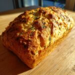 A golden-brown loaf of Italian herb and cheese bread with visible herbs baked into the crust, sitting on a wooden board.