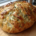 A freshly baked, golden-crusted loaf of Italian herb and cheese bread resting on a wooden cutting board.