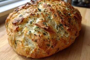 A freshly baked, golden-crusted loaf of Italian herb and cheese bread resting on a wooden cutting board.