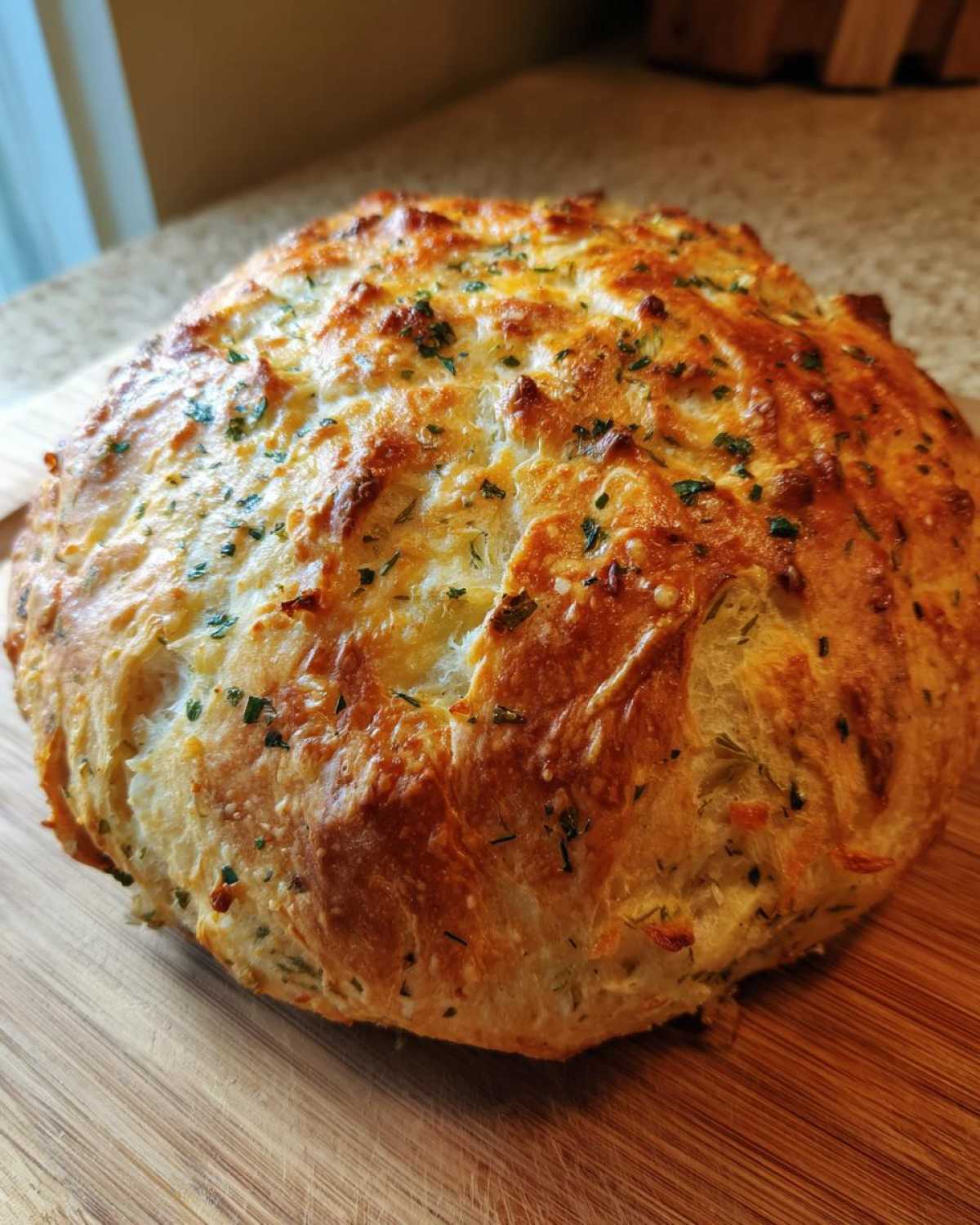 A freshly baked, golden-brown loaf of Italian herb and cheese bread resting on a wooden cutting board.
